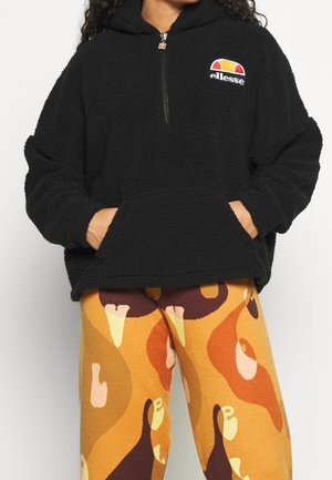 Mustahdollinen musta fleece-huppari, jossa on puolivetoketju ja etutaskut, sekä brodeerattu logo. Yhdistetty kuvioituihin keltaisiin, ruskeisiin ja oransseihin legginsseihin.