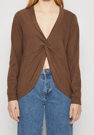 Vrouw draagt een bruine top met lange mouwen en een gedraaide voorkant, gecombineerd met een high-waisted blauwe spijkerbroek, waarbij de buik zichtbaar is en de broek een knoopsluiting heeft.