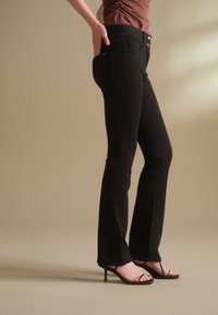Person trägt schwarze, eng anliegende Hose und schwarze Sandalen mit hohen Absätzen, steht auf einem beigefarbenen Boden vor einem beigefarbenen Hintergrund.