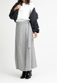 Vrouw in een witte top, grijze wikkelrok met riem, zwarte pufferjack over de schouders en zwarte enkelhakschoenen op een witte achtergrond.