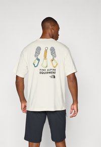 Light beige t-shirt featuring a back graphic of climbing gear: two ropes and three carabiners in various colors. Paired with dark shorts.