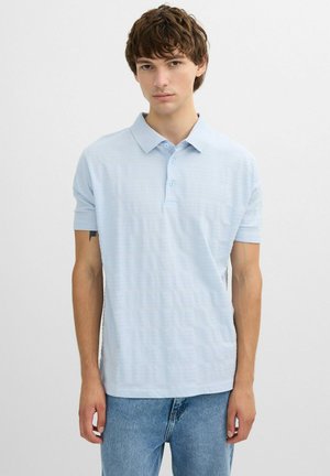 Joven con cabello corto y rizado, vistiendo una camisa polo de manga corta azul claro y jeans azules, de pie sobre un fondo blanco liso.