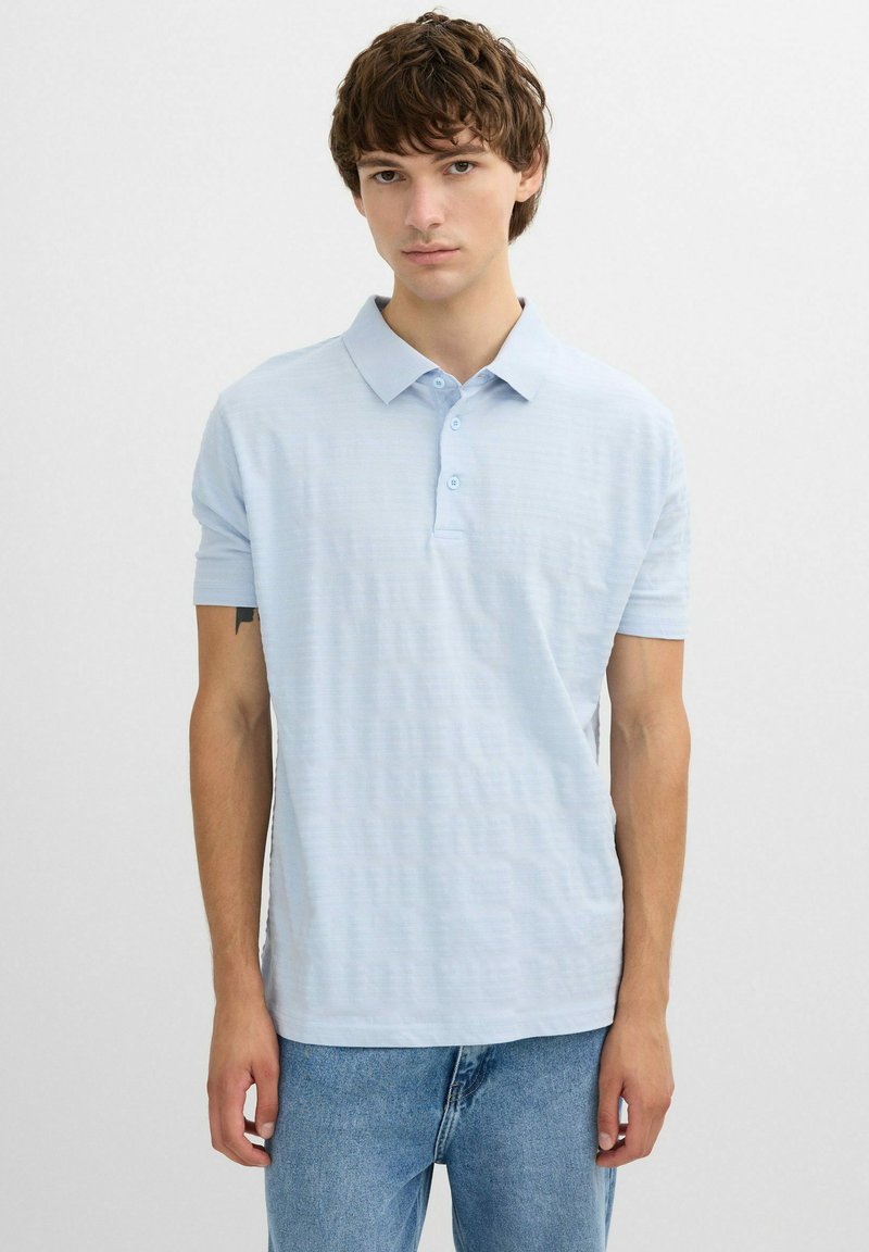 Joven con cabello corto y rizado, vistiendo una camisa polo de manga corta azul claro y jeans azules, de pie sobre un fondo blanco liso.