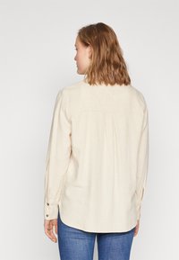 Light beige long-sleeve shirt with a back yoke, pleats, and buttoned cuffs; made of a soft, textured fabric; paired with blue jeans.