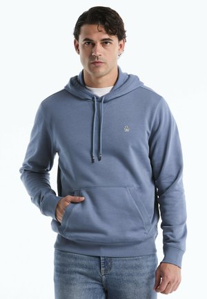 Mann trägt einen blauen Hoodie mit einer Fronttasche und Kordelzügen, kombiniert mit hellblauen Jeans, stehend vor einem weißen Hintergrund.