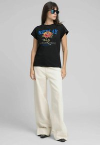 Maglietta nera con scritta blu "REPLAY" e grafica di rosa, abbinata a pantaloni crema a gamba larga e scarpe nere. Occhiali da sole completano il look.