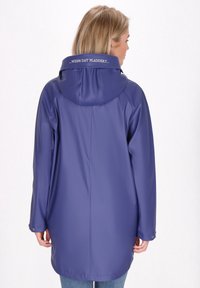 Fialový nepremokavý plášť do dažďa s kapucňou, ktorý má dlhší strih, cvokové uzáveri a textový detail na golieri. Hladký, odolný materiál.
