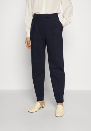 Kobieta nosząca ciemnoniebieskie, zwężane spodnie z wysokim stanem i plisami, w połączeniu z bluzką w kolorze ecru i kremowymi niskimi butami wsuwanymi.