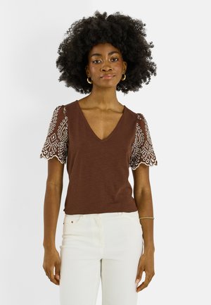 Mujer con peinado afro que lleva una camiseta marrón con cuello en V y mangas cortas de encaje blanco, y pantalones blancos, de pie frente a un fondo blanco.