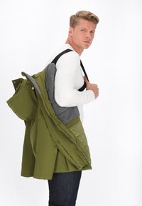 Olivgrön jacka med grå foder, med dragkedjor och fickor, modell håller jackan över axeln, bär en vit långärmad skjorta.