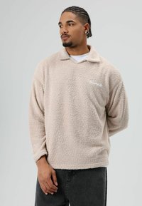 Lichtbeige fleece pullover met een kraag en een wit geborduurd logo. De textuur is zacht en pluizig, met lange mouwen en een losse pasvorm.