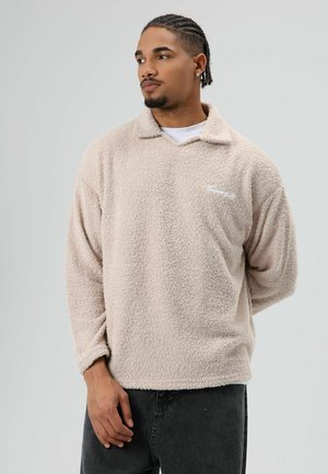 Lichtbeige fleece pullover met een kraag en een wit geborduurd logo. De textuur is zacht en pluizig, met lange mouwen en een losse pasvorm.