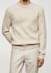 Maglione beige con un motivo geometrico testurizzato, colletto e polsini a coste. Abbinato a pantaloni chiari, con una vestibilità rilassata.