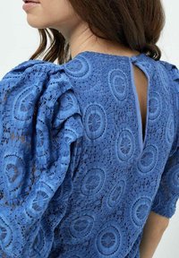 Blusa in pizzo blu con maniche a sbuffo, motivi circolari e dettaglio a chiave sulla schiena. Il materiale presenta un designintricato e testurizzato.