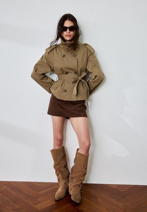 Femme portant une veste beige surdimensionnée ceinturée, une mini-jupe marron foncé, des bottes beige décontractées et des lunettes de soleil noires, debout contre un mur blanc.