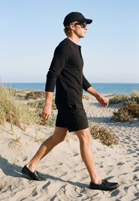 Uomo con berretto nero, occhiali da sole, camicia a maniche lunghe, pantaloncini e scarpe che cammina su una spiaggia sabbiosa con l’oceano sullo sfondo.