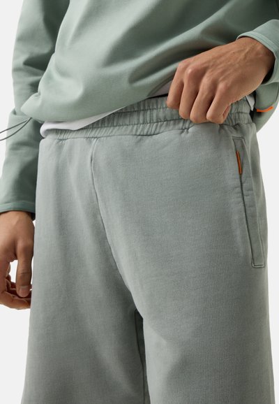 Personne portant un pantalon de survêtement vert clair avec une taille élastique, tirant légèrement sur la taille avec la main, et un sweat-shirt assorti visible.