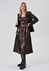 Schwarze Lederjacke über einem blumigen satin Kleid in Bronze und Schwarz, knielang, mit einer ausgestellten Silhouette und klobigen schwarzen Stiefeln bis zum Knöchel.