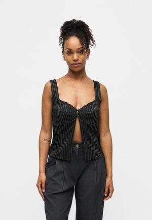 Femme aux cheveux bouclés attachés en queue de cheval haute, portant un haut sans manches noir à fines rayures avec des crochets à l'avant et un pantalon plissé gris foncé.