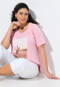 Lyserød t-shirt med korte ærmer, der har et grafisk print af et ørkenlandskab med kaktusser og en sol. Sammen med hvide, knælange shorts.