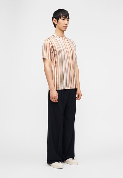 Άνδρας που στέκεται, φοράει κοντομάνικο πουκάμισο με πολύχρωμες κάθετες ρίγες, φαρδιά μαύρα παντελόνια και λευκά αθλητικά παπούτσια, μπροστά σε λευκό φόντο.