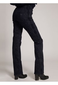 Jeans blu scuro con una stampa a serpente testurizzata, caratterizzati da un design aderente, vita media e gambe svasate, abbinati a stivali neri con tacco.
