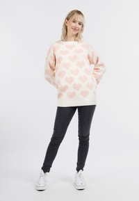 Maglione lavorato a maglia di colore crema con motivi a cuore rosa chiaro, caratterizzato da una vestibilità ampia, maniche lunghe e orlo a coste. Abbinato a jeans skinny neri.