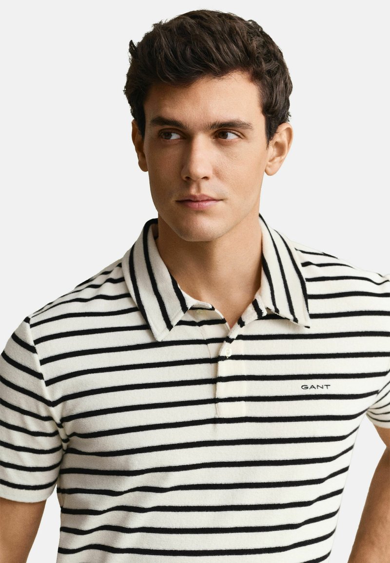 Jeune homme aux cheveux courts et foncés portant un polo blanc avec des rayures horizontales noires et un petit logo "GANT" sur la poitrine.