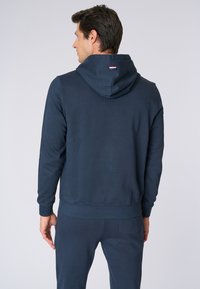 Felpa blu navy realizzata in tessuto morbido, con cappuccio regolabile tramite stringa e polsini a coste. Comprende un piccolo dettaglio a forma di bandiera sul retro.