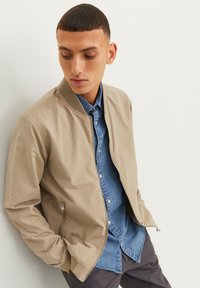 Veste bomber beige avec une fermeture éclair et des poignets côtelés, portée par-dessus une chemise en denim. Tissu lisse avec une finition mate, design simple.