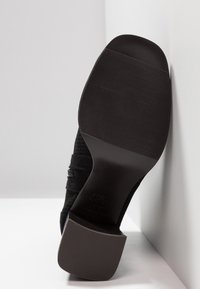 Chaussure à talons hauts noire avec une semelle lisse et texturée. Comprend un design de bride et une large base de talon. Logo de la marque visible sur la semelle.