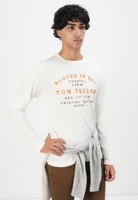 Jonge man in een wit T-shirt met lange mouwen met oranje en zwarte tekst, bruine broek en een grijze trui om zijn middel geknoopt.