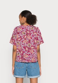 Blusa às mangas curtas em cor bordô com padrões de paisley e florais. Tecido suave, corte relaxado, decote redondo e detalhes de costura visíveis.