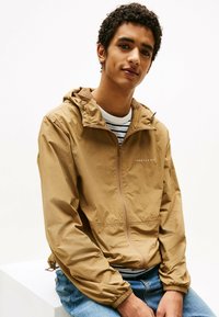 Chaqueta ligera color beige con cremallera frontal y capucha, puños elásticos y un logotipo bordado. Lleva puesta sobre una camiseta a rayas y unos jeans azules.