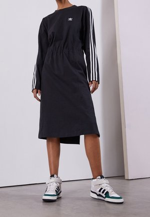 Osoba ubrana w czarną sukienkę Adidas z białymi paskami na rękawach oraz białe sportowe trampki Adidas na wysokim kroku z czarnymi i zielonymi akcentami.