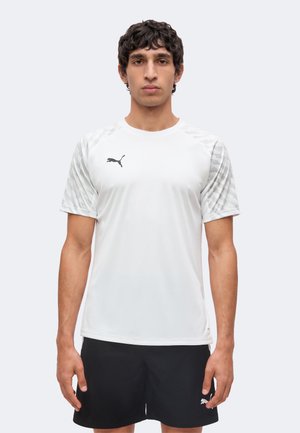 Mand står med fronten fremad, iført en hvid Puma sportstrøje med mønstrede ærmer og sorte shorts mod en ensfarvet lys baggrund.