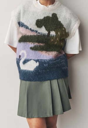 Osoba nosząca puszystą kamizelkę swetrową z pejzażowym wzorem przedstawiającym białego łabędzia, drzewo i wodę, w połączeniu z białą koszulą i zieloną plisowaną spódnicą.