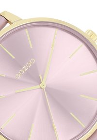Montre ronde avec un cadran texturé rose clair, des index et aiguilles dorés, et un boîtier doré. Le nom de la marque "oozoo" est affiché au centre.