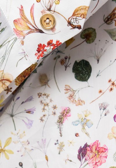 Fehér anyag virágmintával, színes virágokkal és növényekkel, aranygombbal díszítve a gallérnál.