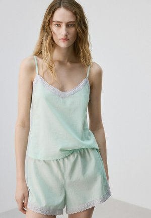 Giovane donna con capelli mossi, indossa una canottiera verde menta chiaro e pantaloncini coordinati con rifiniture in pizzo bianco, in piedi davanti a uno sfondo neutro.