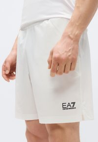 Biele športové šortky s hladkou textúrou, elastickým pásom, bočnými vreckami a logom EA7 Emporio Armani na ľavej nohe.