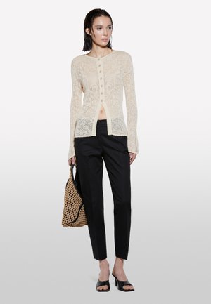 Cardigan en dentelle couleur crème avec un col rond, fermeture à boutons et ourlet fendu. Associé à un pantalon slim noir et un sac en tissé.