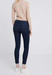 Mörkblå tajta jeans med hög midja, föreslår bakfickor och en diskret logotypdetalj. Kombinerat med en ljusbeige ribbad tröja och vita sneakers.