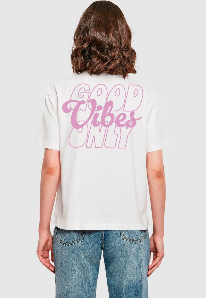 Valge puuvillane t-särk, millel on seljal suur roosa graafiline tekst "GOOD VIBES ONLY", lõdva istuvuse ja lühikeste varrukatega.