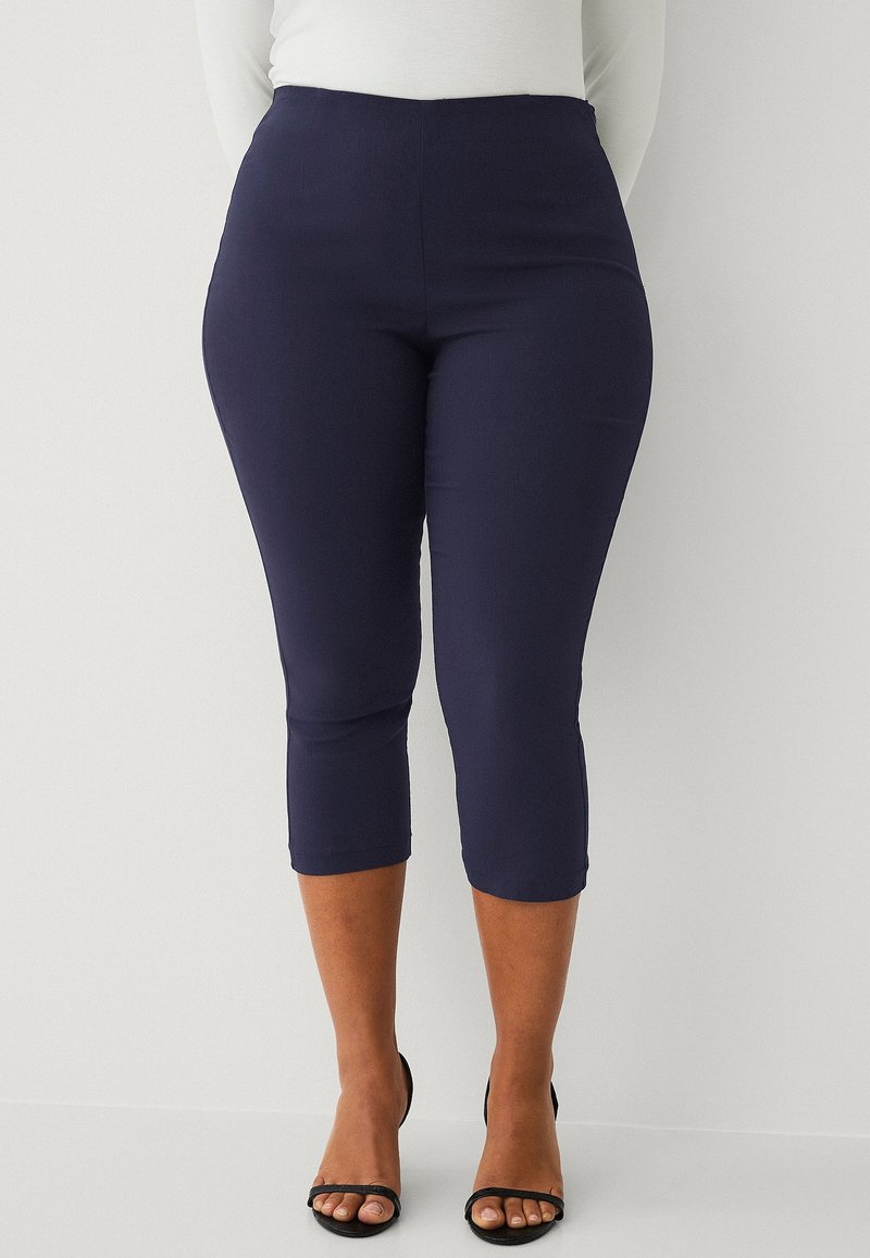 Pantaloni capri blu navy realizzati in materiale elastico, con vita alta e taglio alla caviglia, abbinati a tacchi aperti neri.