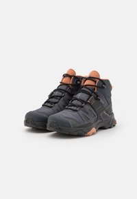 Scarpe outdoor alte realizzate in tessuto grigio scuro con accenti arancioni e suole in gomma nera, dotate di cuciture dettagliate e una punta rinforzata.