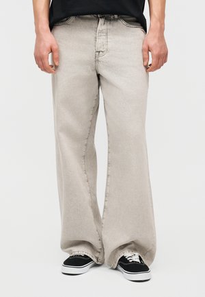 Άτομο που φορά ανοιχτό γκρι τζιν με φαρδιά παντελόνια, συνδυασμένο με μαύρα και λευκά αθλητικά παπούτσια, στέκεται μπροστά σε απλό φόντο.