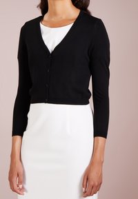 Czarny sweter z dzianiny z dekoltem w serek, długimi rękawami i pięcioma guzikami z przodu. Nosi się go na białej, fakturowanej sukience.