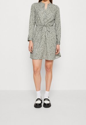 Femme portant une robe verte à manches longues avec des boutons et des taches blanches, nouée à la taille, associée à des chaussettes blanches et des mocassins noirs.