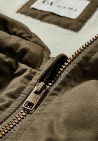 Fermeture éclair en bronze sur une veste en tissu vert olive, présentant une texture matelassée et une étiquette en tissu blanc avec la mention "64 MAMA". Intérieur en matériau doux visible.